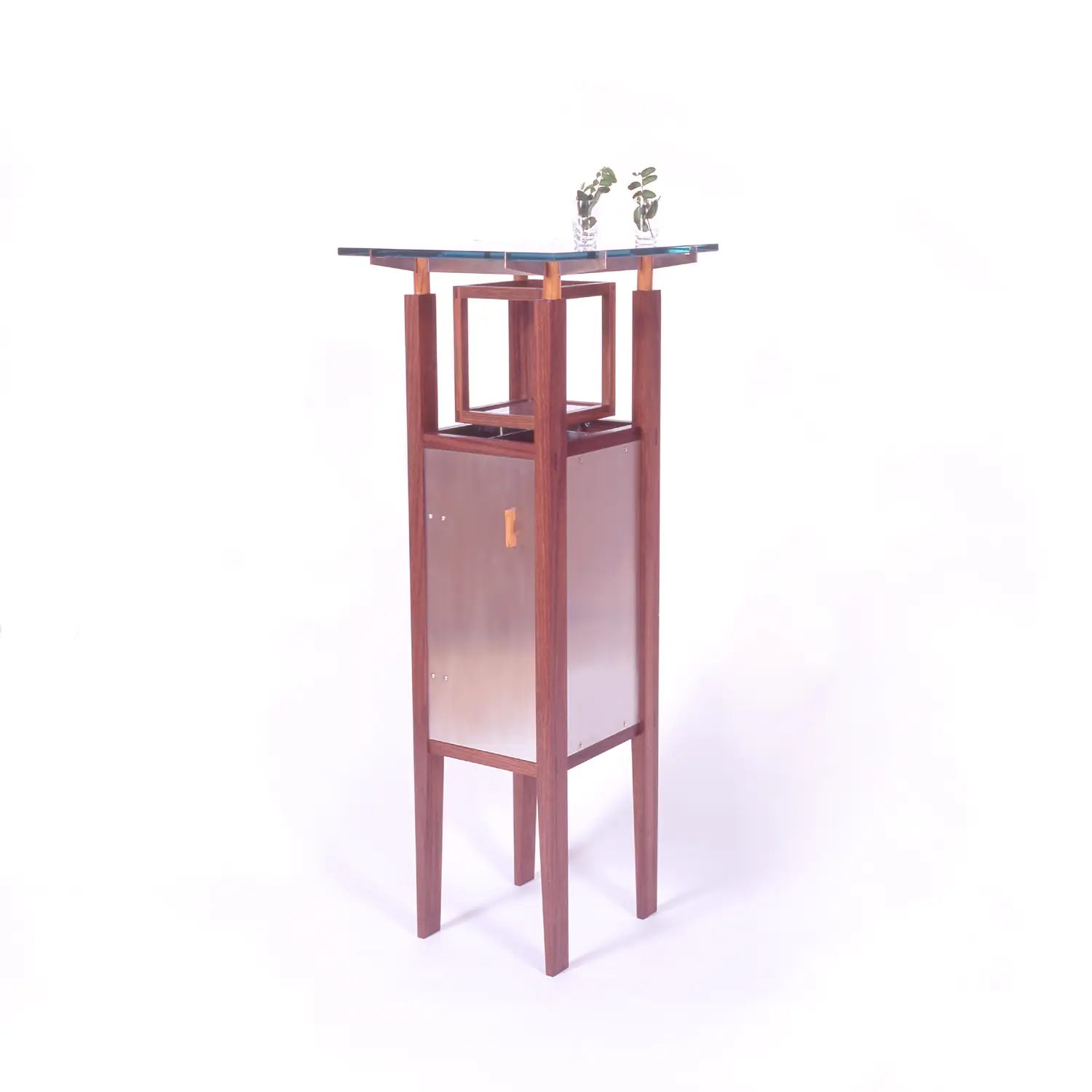 Isan Table with a tall walnut framework, contrasting metal cabinet, and transparent glass top